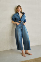 Eine Frau ist lässig an eine Wand angelehnt und trägt ein Jeanshemd mit weißen Streifen 27_THE SUIT UP BLOUSE STRIPES in mittelblau und dazu die passenden Jeans mit weißen Streifen 05_THE O SHAPE EXTREME STRIPES in mittelblau.