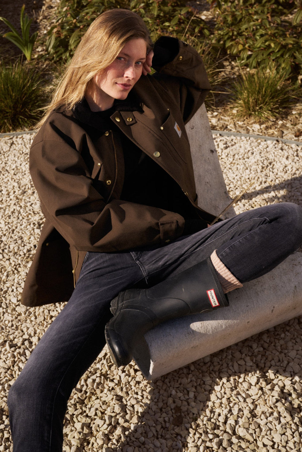 Eine Frau sitzt draußen auf einem Betonstuhl und hat ihr linkes Bein angewinkelt. Ihr linker Arm ist auf der Rückenlehne abgestützt. Sie trägt die 02_THE SLIM in dunkelgrau mit Gummistiefeln.