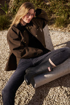 Eine Frau sitzt draußen auf einem Betonstuhl und hat ihr linkes Bein angewinkelt. Ihr linker Arm ist auf der Rückenlehne abgestützt. Sie trägt die 02_THE SLIM in dunkelgrau mit Gummistiefeln.