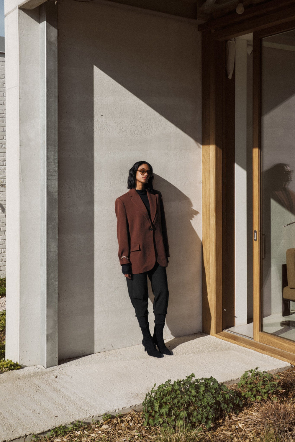 Eine Frau ist an eine Hauswand angelehnt. Die Sonne scheint auf sie. Sie trägt eine Sonnenbrille und einen braunen Blazer, dazu hat sie eine Hose 06_THE JINO O JOGGER in schwarz kombiniert.