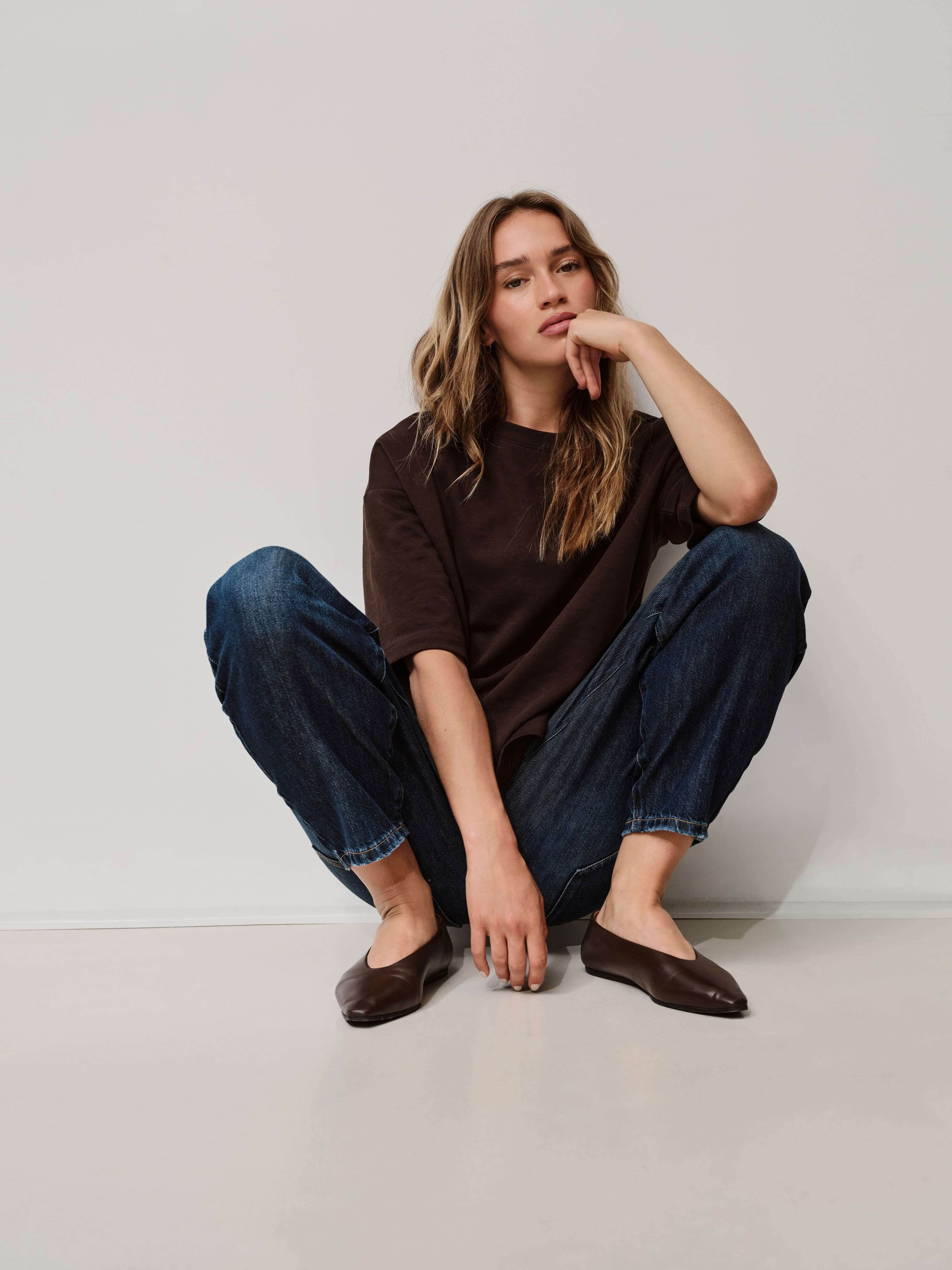 Frau in braunem T-Shirt und dunkler Jeans, hockend vor einer hellen Wand.