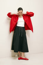 Frau in sportlicher roter Jacke, schwarzem Midi-Rock und roten Ballerinas, posierend vor hellem Studiohintergrund.