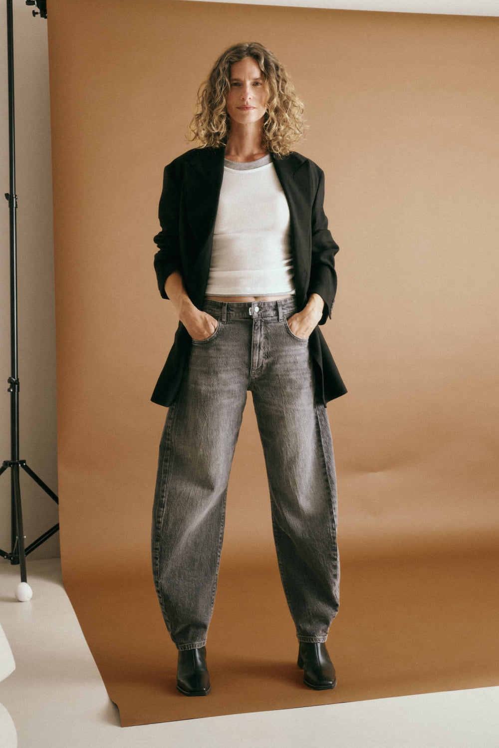 Eine Frau steht in einem Fotostudio vor einer rostbraunen Leinwand. Sie trägt ein weißes T-Shirt mit einem schwarzen Blazer darüber, dazu eine Jeans 05_THE O SHAPE LONG in mittelgrau.