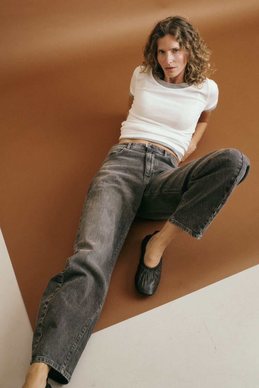 Eine Frau liegt lässig auf dem Boden, der Hintergrund ist rostbraun. Sie trägt ein weißes T-Shirt und dazu die Jeans 05_THE O SHAPE LONG in mittelgrau.