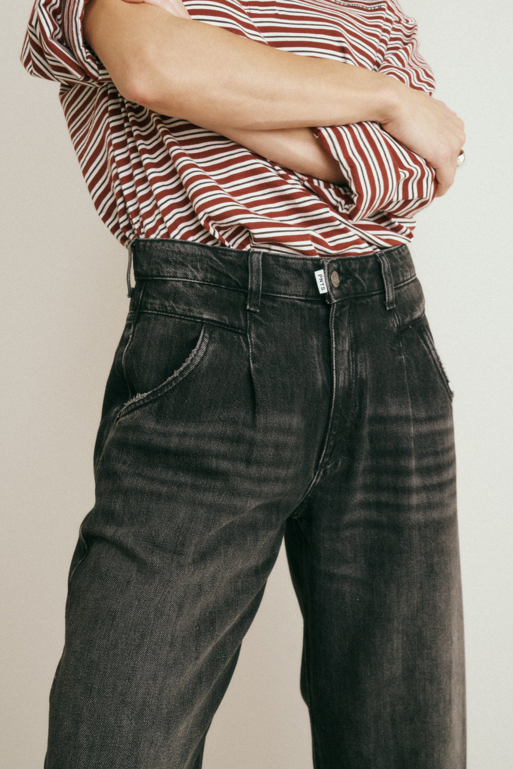 Detailansicht: Man sieht die eingearbeiteten Bundfalten vorn an der Jeans 05_THE O SHAPE PLEAT in dunkelgrau sowie ein PNTS-Fähnchen an der Knopfleiste.