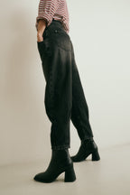 Detailansicht: Man sieht die seitliche Ansicht der Jeans 05_THE O SHAPE PLEAT in dunkelgrau. Hinten am Saum sind Kellerfalten sichtbar. Dazu trägt sie schwarze Stiefel.