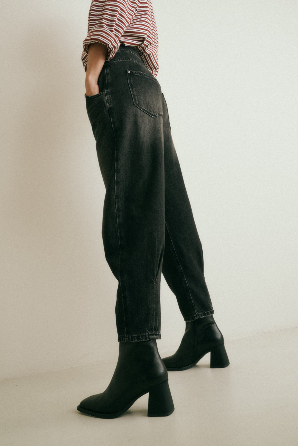 Detailansicht: Man sieht die seitliche Ansicht der Jeans 05_THE O SHAPE PLEAT in dunkelgrau. Hinten am Saum sind Kellerfalten sichtbar. Dazu trägt sie schwarze Stiefel.