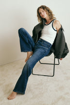 Frau sitzt auf einem Stuhl. Ihre rechtes Bein ist auf dem Stuhl aufgestellt. Sie trägt eine dunkle Jeans, ein weißes Top und eine graue Jacke. Sie schaut in die Kamera.