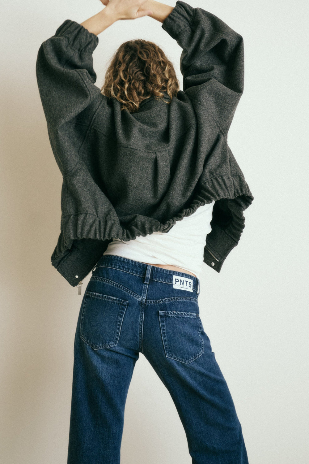 Frau steht vor einer Wand. Man sieht sie von hinten. Ihre Arme hat sie in der Luft. Sie trägt eine dunkelblaue Jeans, ein weißes Top und eine graue Bomberjacke.