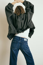 Frau steht vor einer Wand. Man sieht sie von hinten. Ihre Arme hat sie in der Luft. Sie trägt eine dunkelblaue Jeans, ein weißes Top und eine graue Bomberjacke.