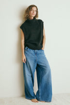 Eine Frau steht vor einer weißen Wand und trägt einen Pullover mit einer lässigen Jeans 22_OVER THE TOP in hellblau.