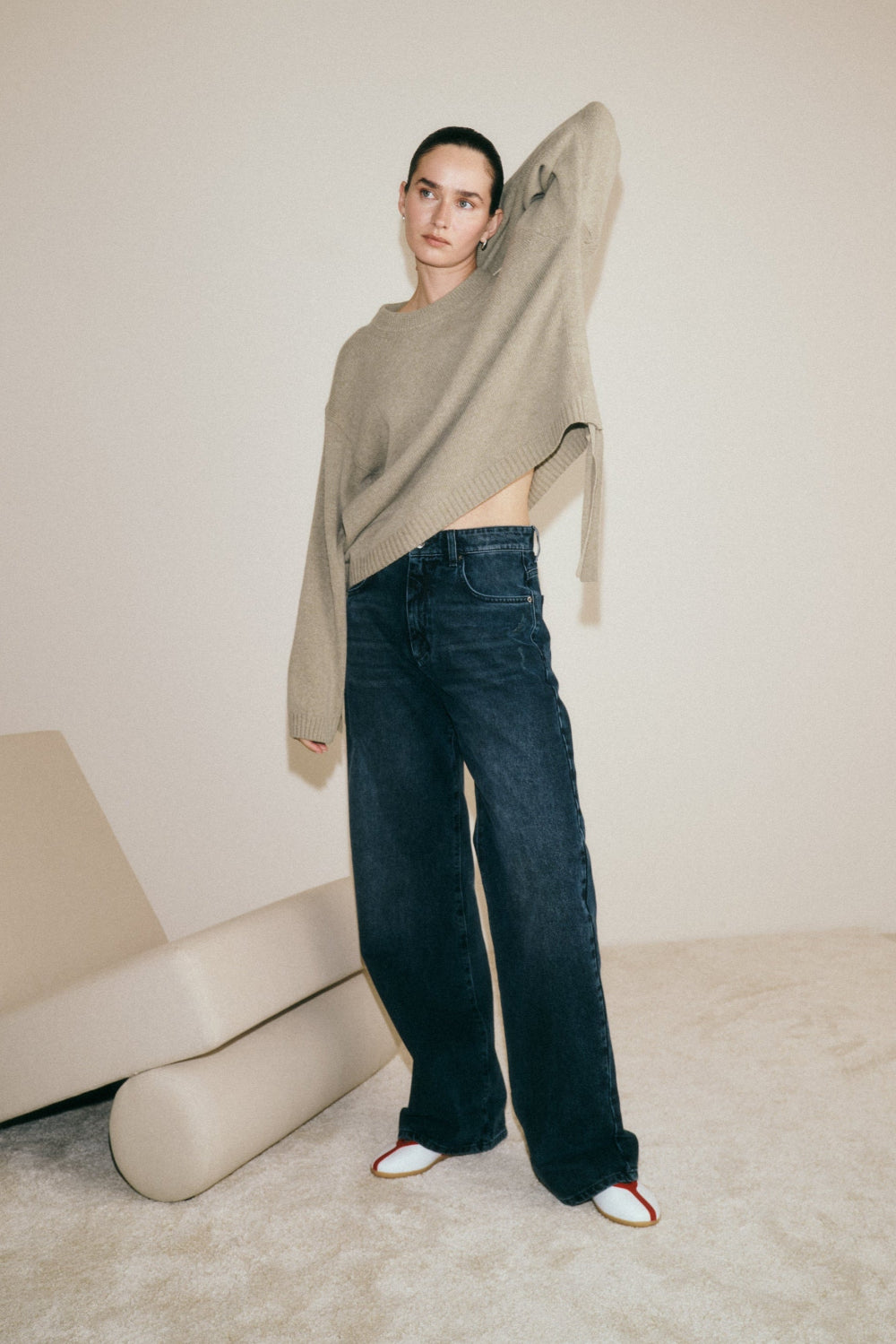 Eine Frau steht vor einer weißen Wand. Sie trägt ein beigen Pullover und dazu die Jeans 26_THE LOOSE in dunkelblau. 