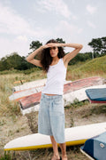 Frau in weißem Ripp-Tanktop und hellblauer Denim-Culotte posiert vor einer Küstenlandschaft mit Booten.