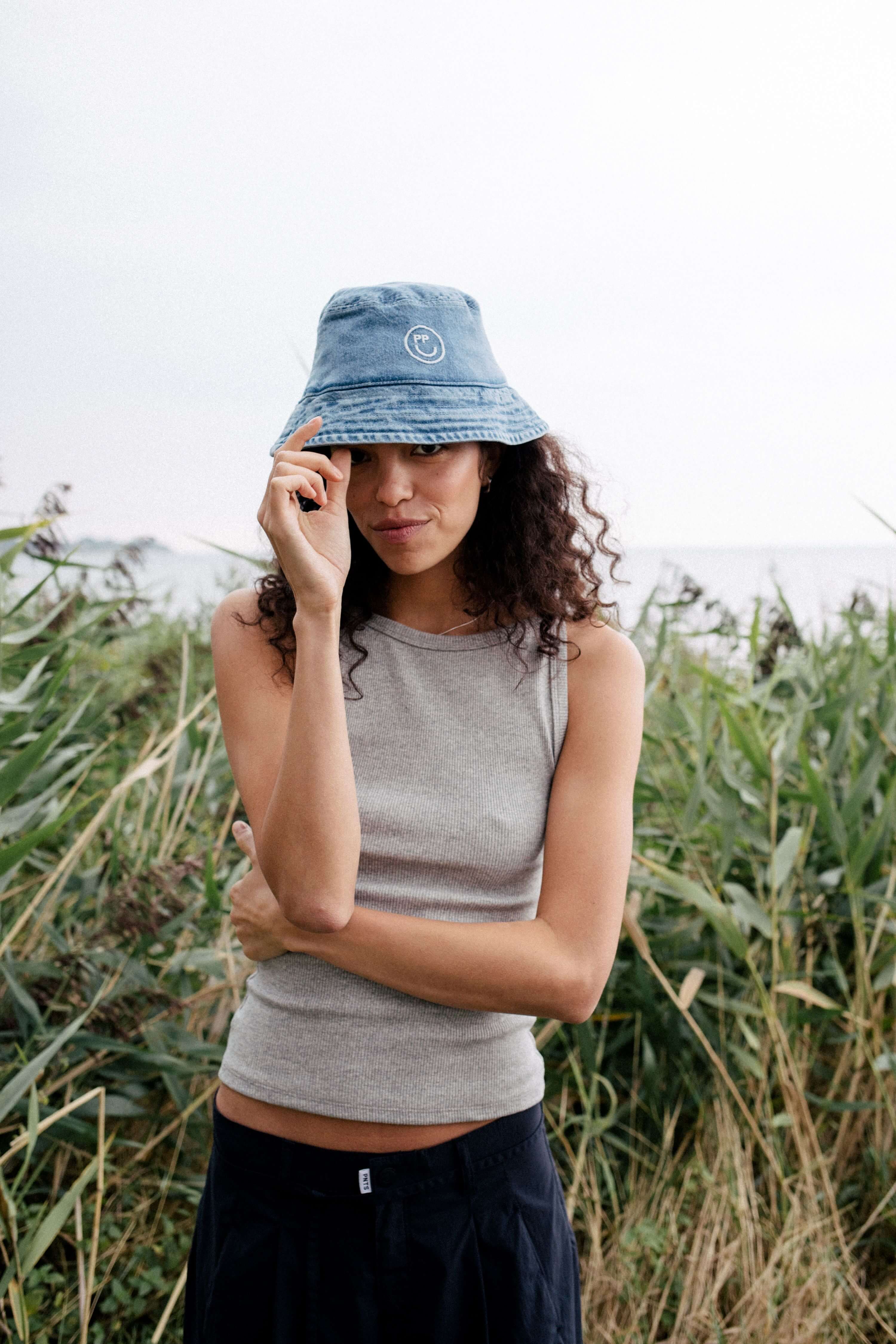 Frau in grauem Ripp-Tanktop trägt einen blauen Denim-Bucket-Hat mit Smiley-Logo vor hohem Schilfgras.