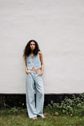 Frau in hellblauem Denim-Set aus Weste und weiter Bundfaltenhose vor einer schlichten weißen Wand.