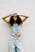 Lachende Frau in hellblauer Denim-Weste und passender weiter Bundfaltenhose vor einer schlichten weißen Wand.