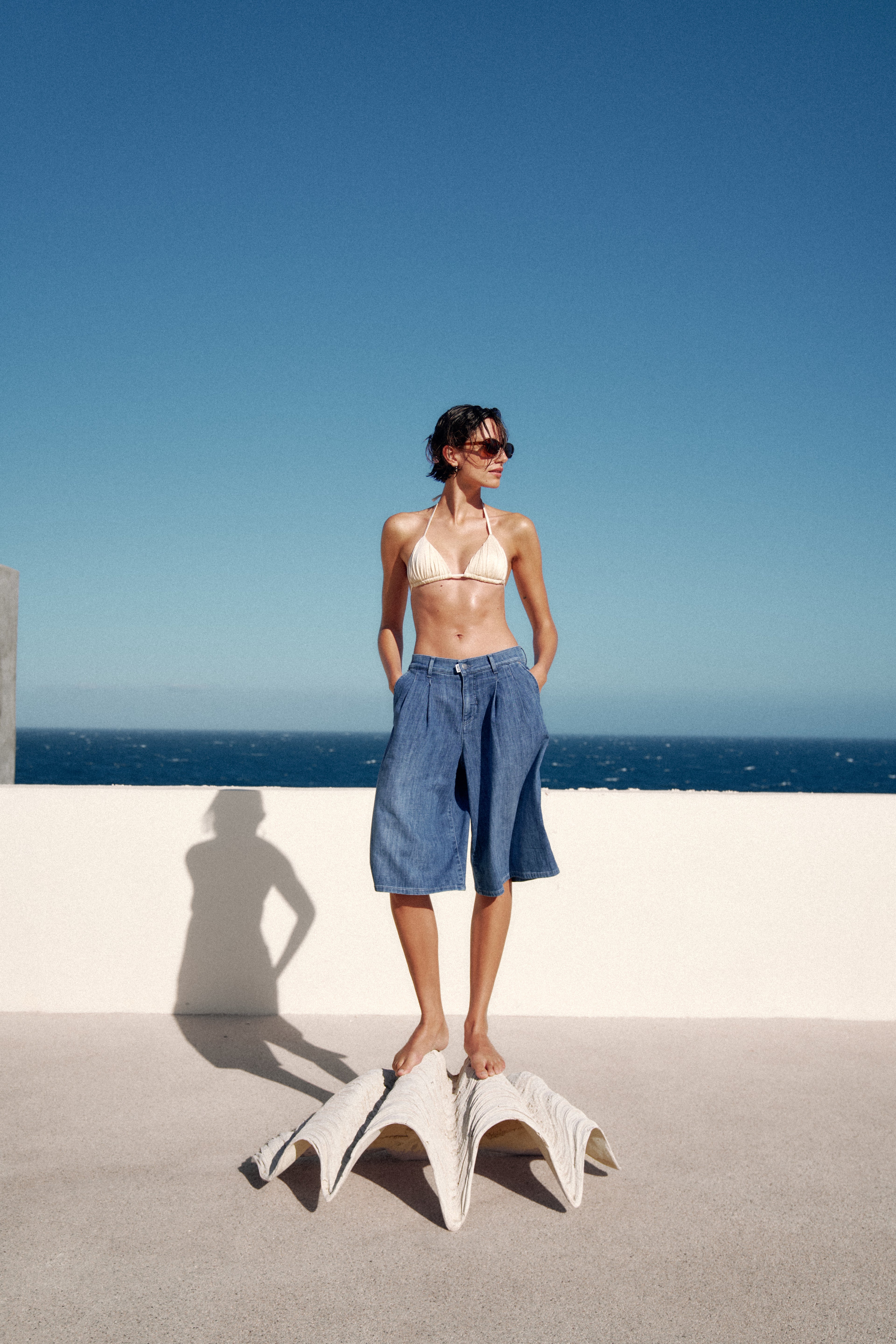 Eine Frau steht auf einer Muschel. Sie trägt ein Bikinioberteil und einer Jeansshorts mit Bundfalte, die kurz bis über das Knie geht. Sie hat die Hände in die Hosentasche gesteckt und schaut nach links. Im Hintergrund sieht man eine hüfthohe Mauer, das Meer und den Himmel.
