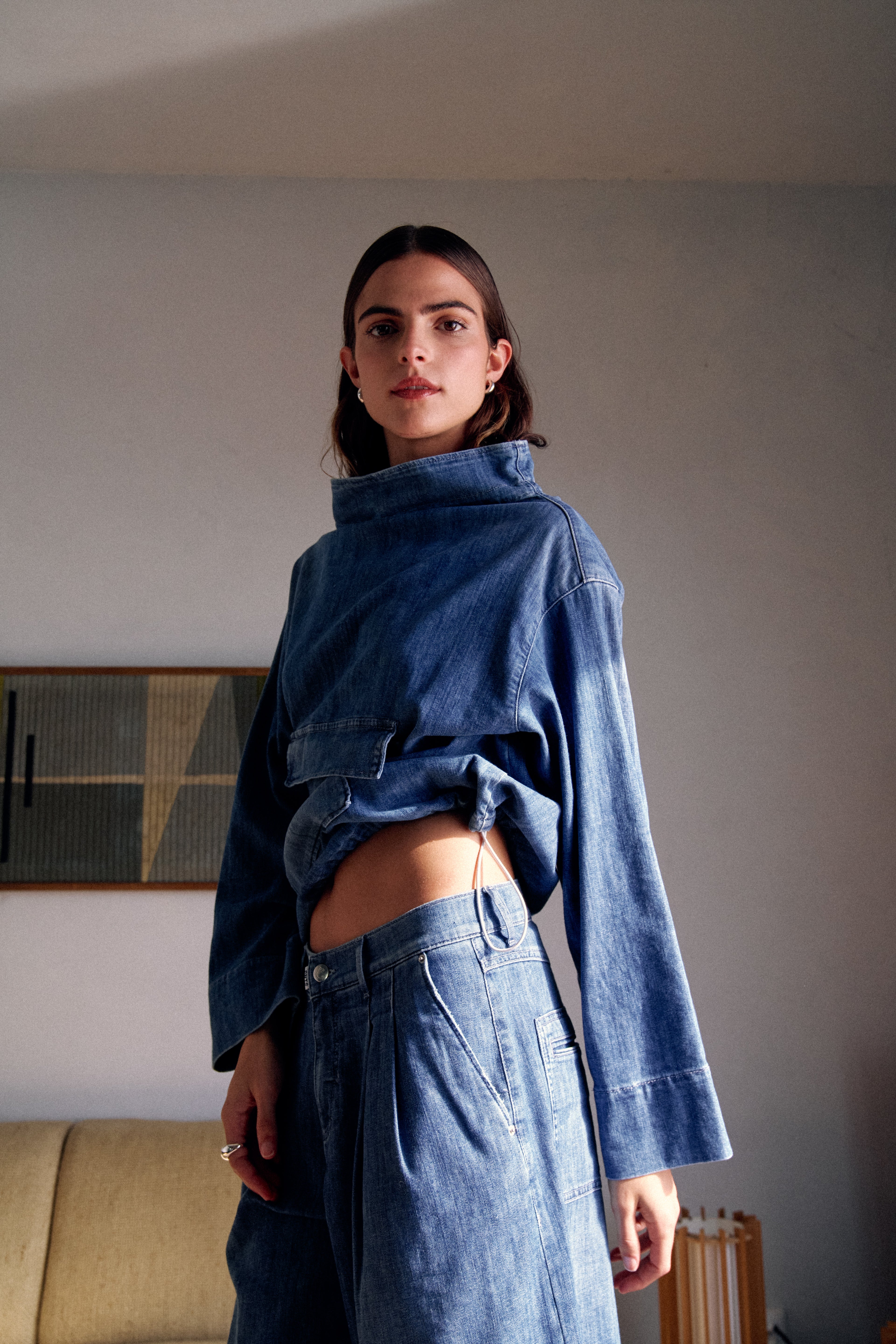 Eine Frau steht in deinem Wohnzimmer. Sie trägt ein Denim Set, das aus einem Denim Pullover und einer Denim Shorts besteht.