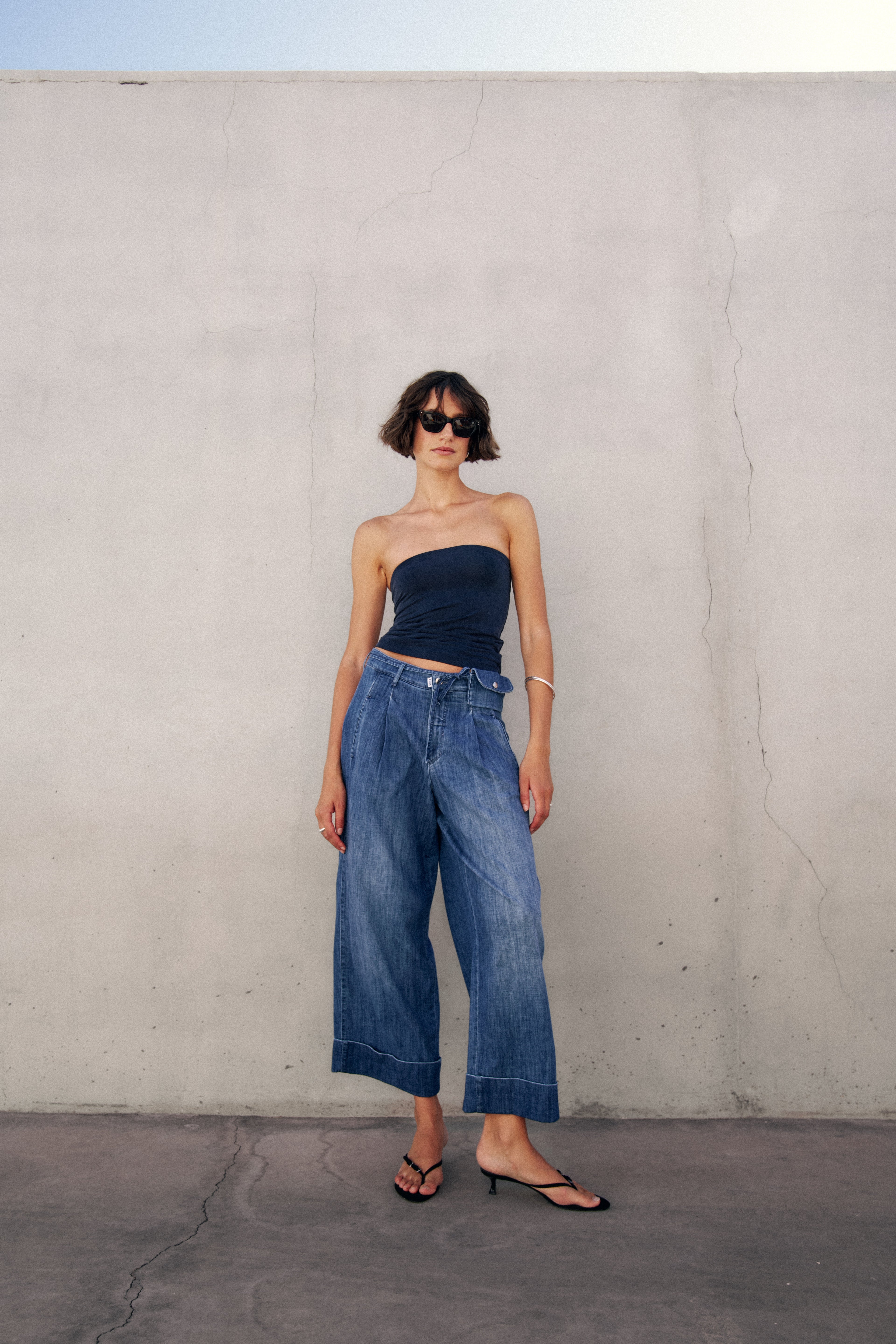 Eine Frau steht vor einer Wand. Sie trägt eine weite, leicht verkürzte Jeanshose, ein dunkelblaues Top und schwarze Flipflops mit Absatz. Sie schaut in die Kamera.