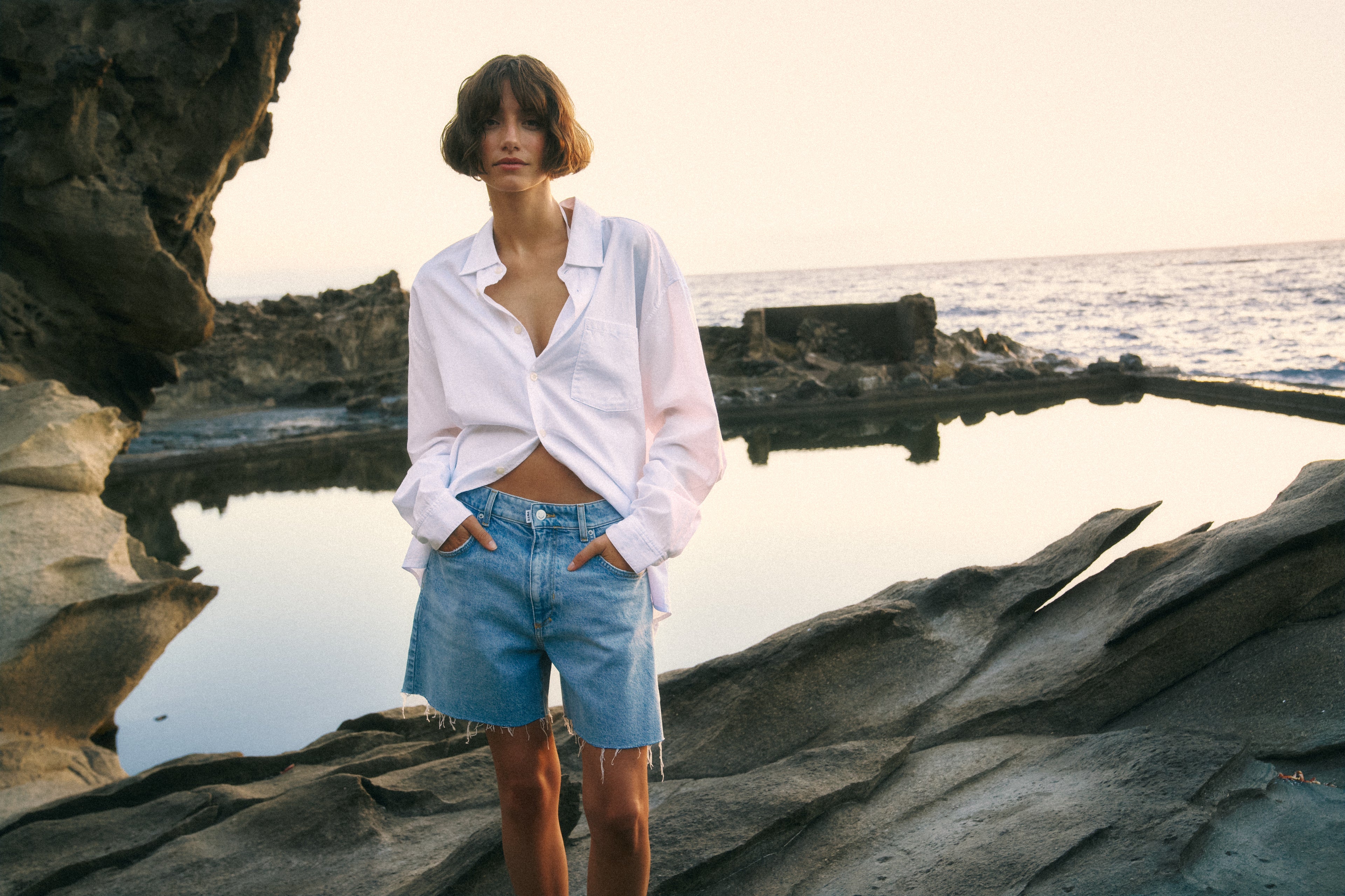 Eine Frau steht auf einem kleinen Fels. Im Hintergrund sieht man das Meer. Sie trägt eine weiße Bluse und eine kurze Jeansshorts. Ihre Hände sind in die Taschen gesteckt.