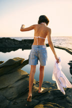 Eine Frau geht über einen kleinen Fels. Man sieht sie von hinten. Sie trägt ein weißes Bikinioberteil und eine kurze Jeansshorts. In ihrer Hand hält sie eine weiße Bluse.