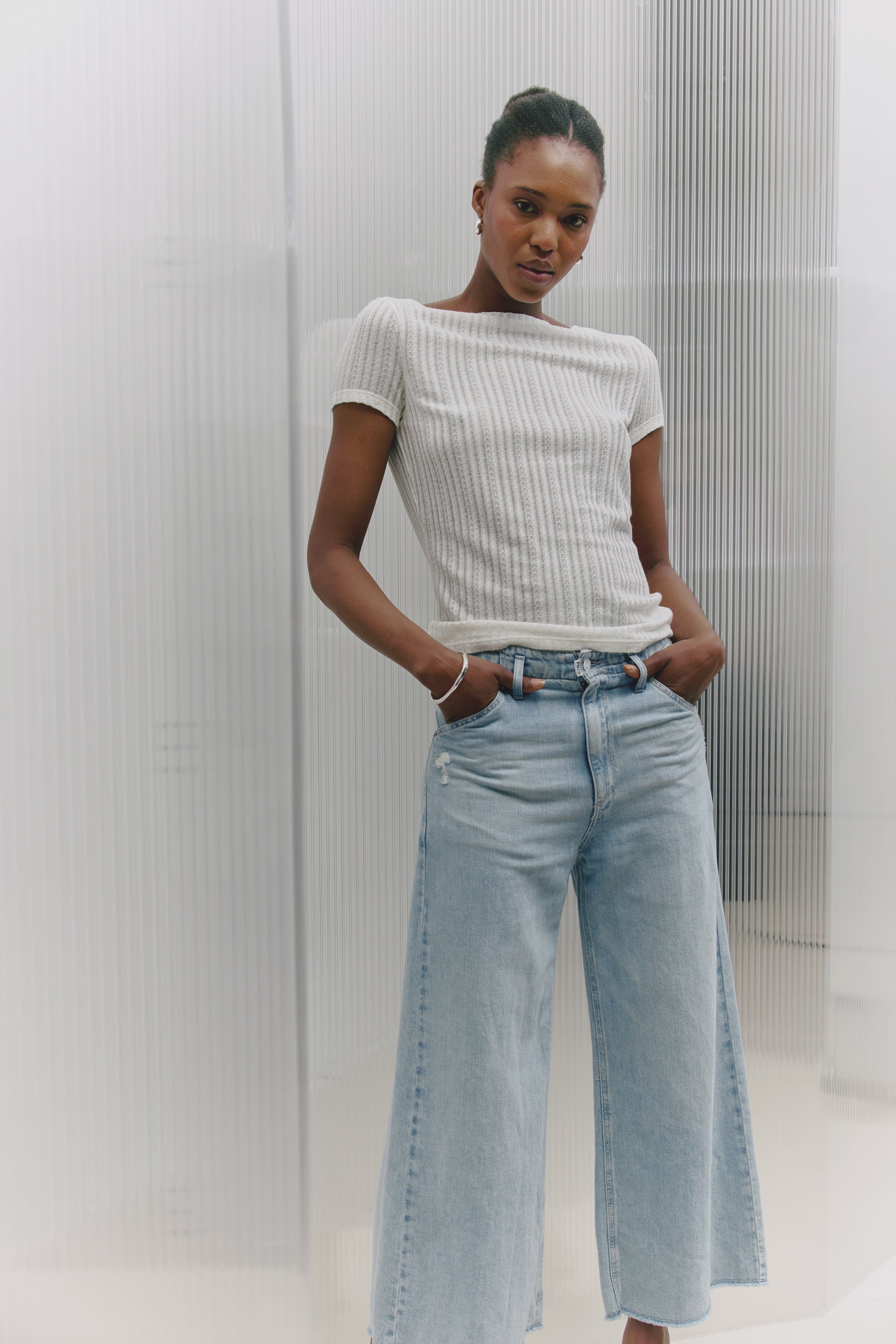 Eine Frau steht vor einer leicht durchsichtigen Wand. Sie schaut in die Kamera und trägt ein weißes T-Shirt mit einer hellblauen, weiten und leicht verkürzten Jeans.