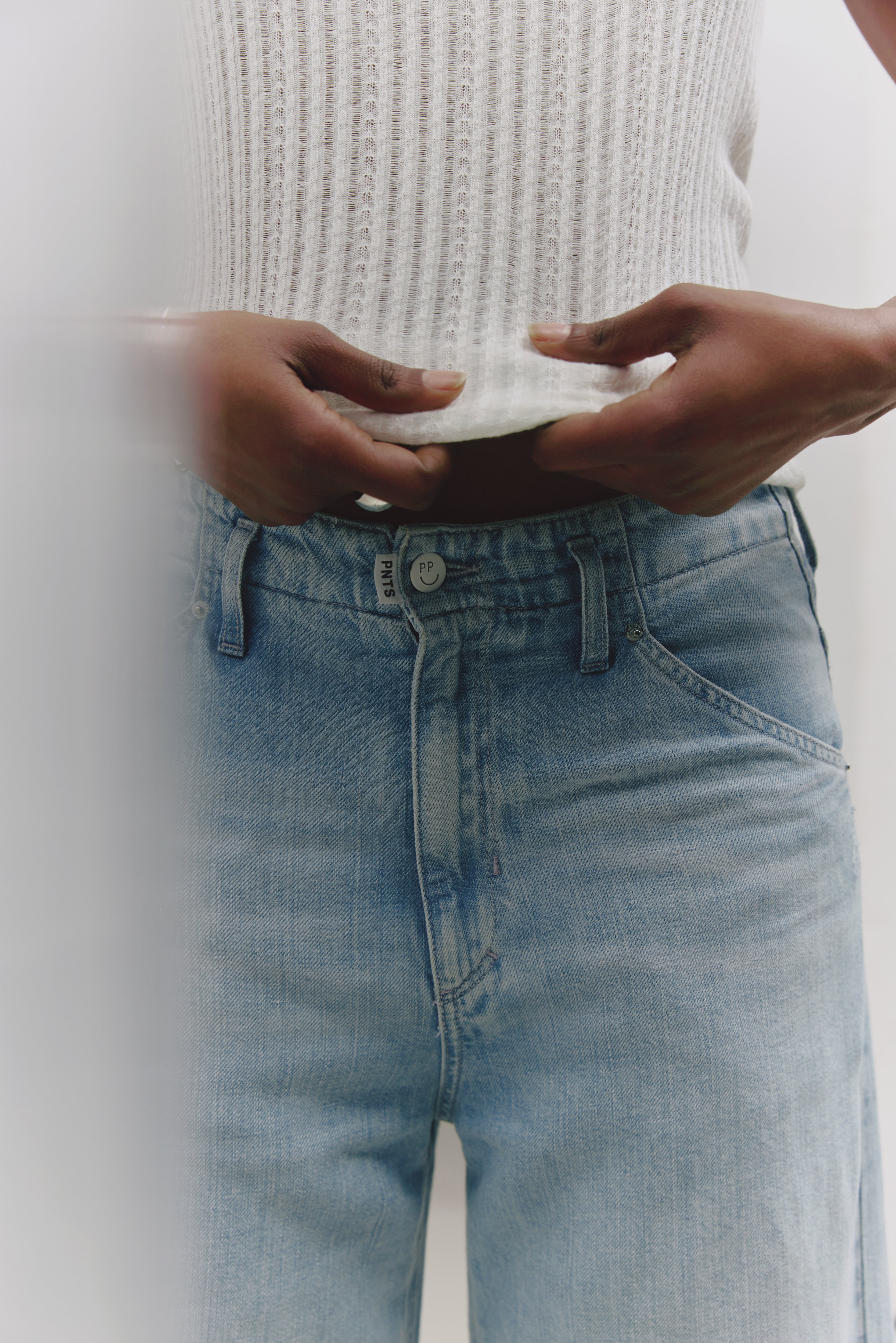 Ein Detailfoto von einer hellen Jeans. Sichtbar ist der PNTS Knopf und das Fähnchen an der Hose. 