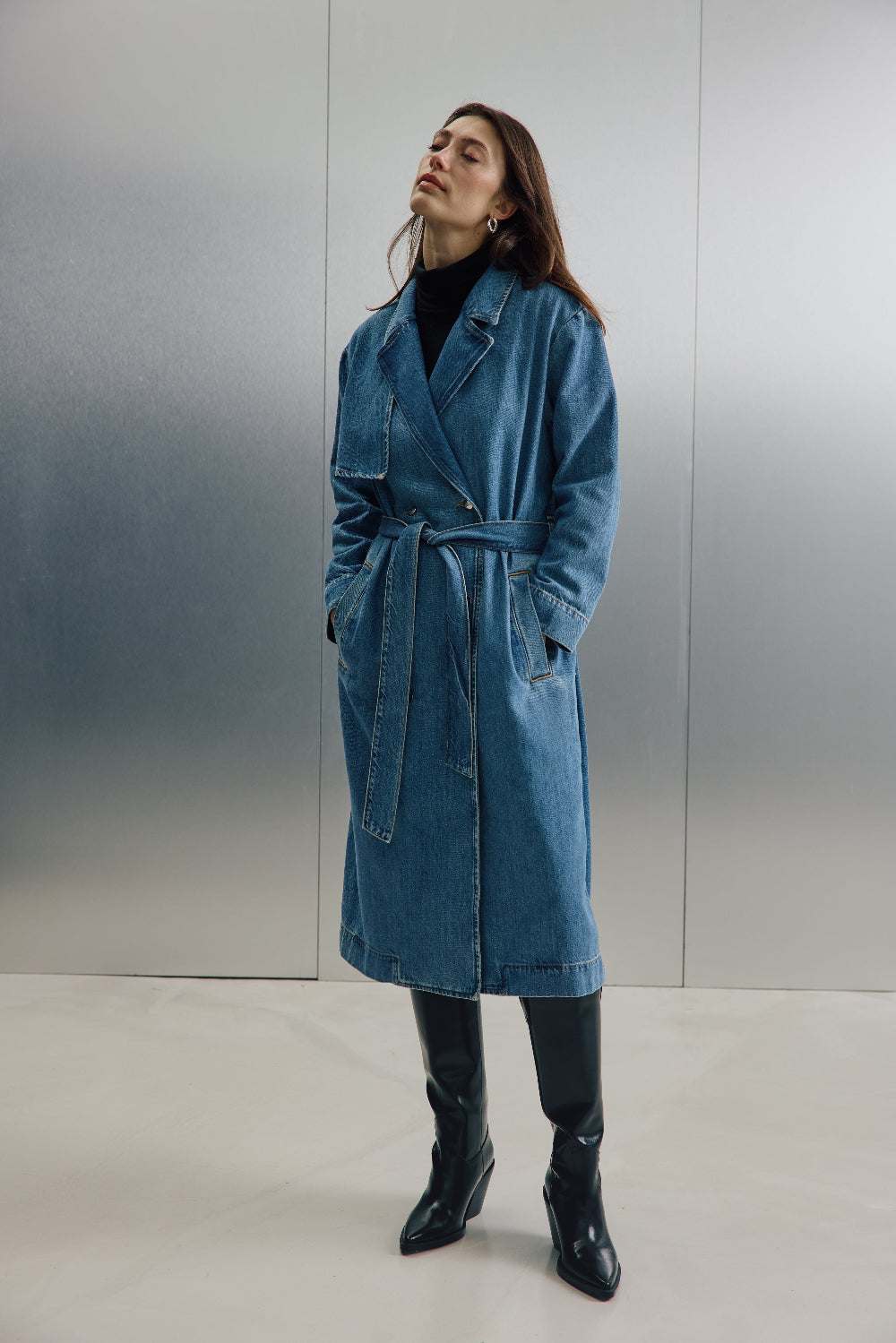 Eine Frau steht vor einer silbernen Wand mit schwarzen Stiefeln. Sie trägt einen geschlossenen Jeans-Trenchcoat 27_THE SUIT UP TRENCH in hellblau.