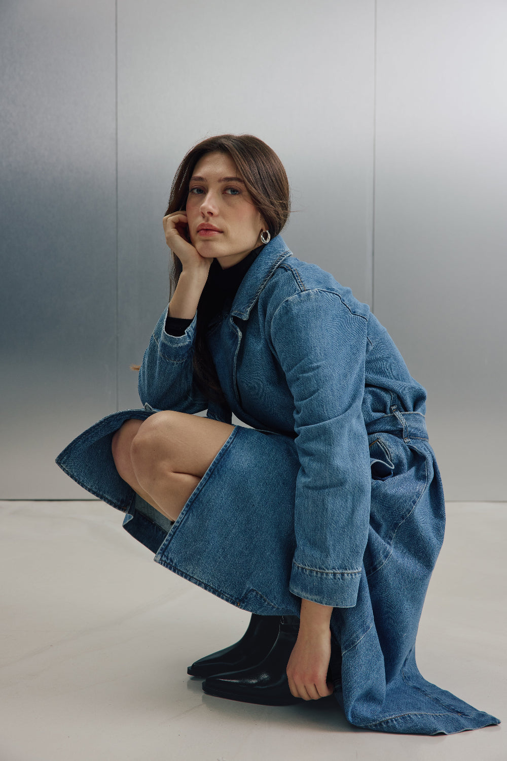 Eine Frau hockt seitlich vor einer silbernen Wand. Sie trägt einen Jeans-Trenchcoat 27_THE SUIT UP TRENCH in hellblau.