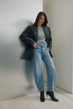 Eine Frau steht angelehnt an einer silbernen Wand. Sie trägt einen hellgrauen Pulli mit einem dunkelgrauen Blazer darüber. Dazu kombiniert sie eine Jeans 05_THE O SHAPE LONG in hellblau.