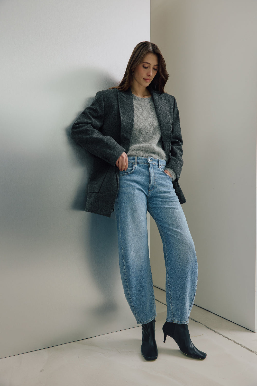 Eine Frau steht angelehnt an einer silbernen Wand. Sie trägt einen hellgrauen Pulli mit einem dunkelgrauen Blazer darüber. Dazu kombiniert sie eine Jeans 05_THE O SHAPE LONG in hellblau.