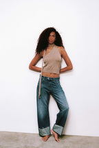 Frau in beige farbenem Neckholder-Top und blauer Jeans mit breitem Umschlag vor weißem Hintergrund.