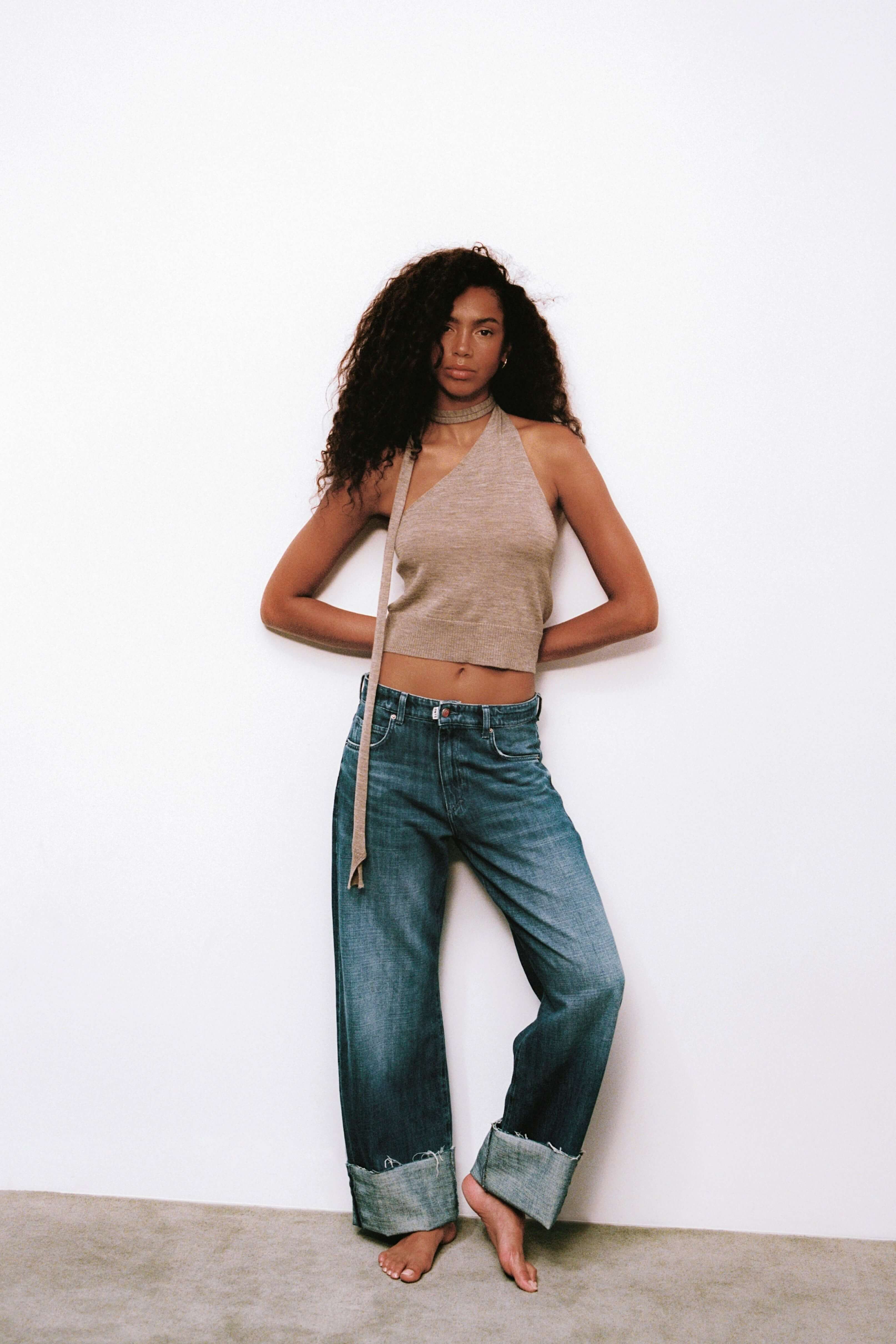 Frau in beige farbenem Neckholder-Top und blauer Jeans mit breitem Umschlag vor weißem Hintergrund.